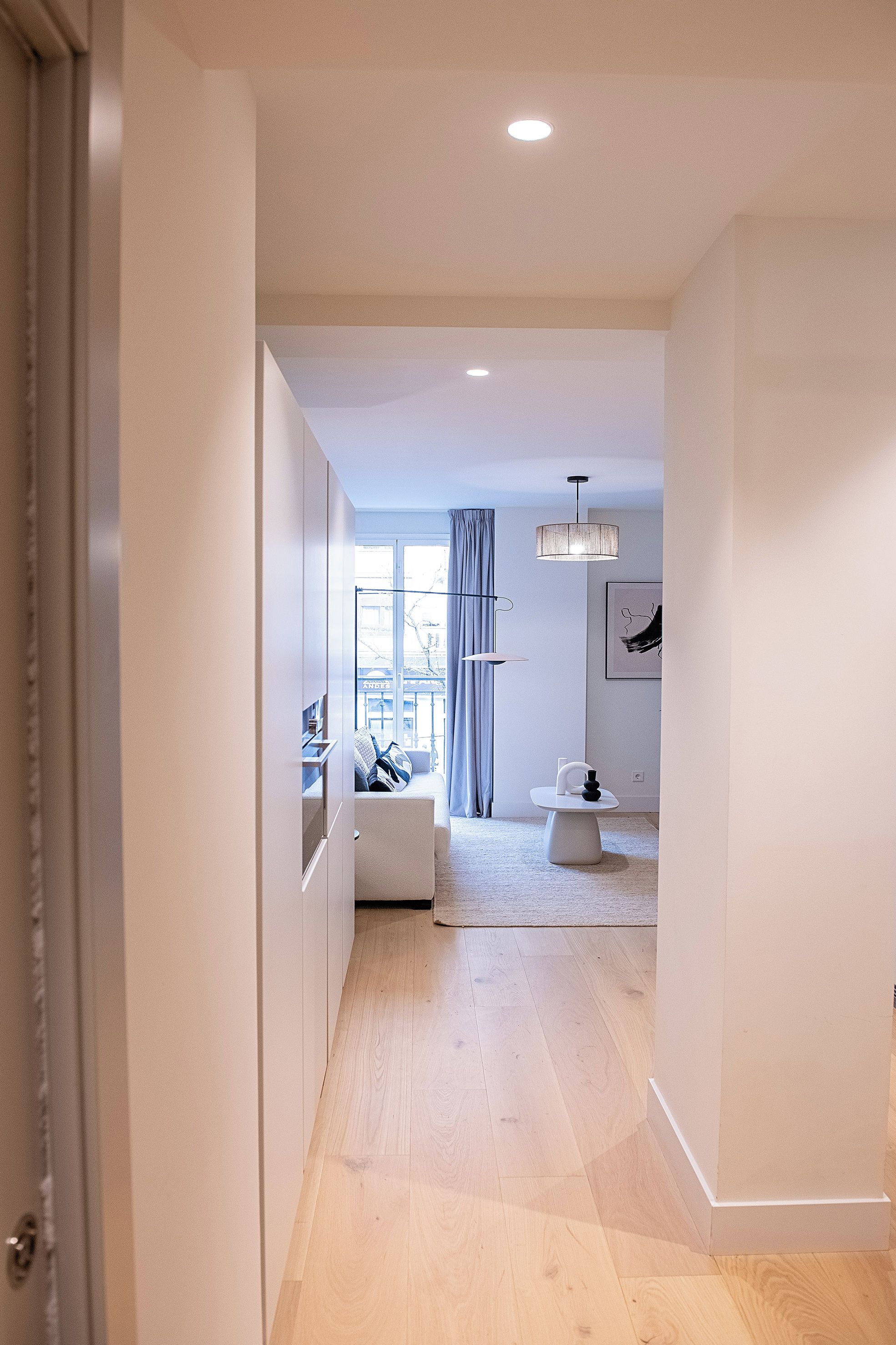 A modern interior with a hallway leading to a living area, featuring a sofa, a table, and a hanging light fixture.
