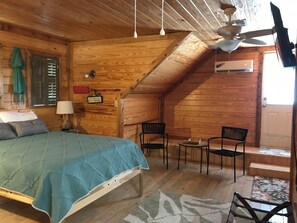 Ein gemütliches Holzzimmer mit einem Bett, zwei Stühlen, einem kleinen Tisch, einer Lampe und einem Deckenventilator.