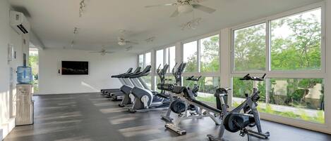 Ein Fitnessraum mit Trainingsgeräten, großen Fenstern, Deckenventilatoren und einem Wasserspender.