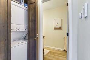 Ein Waschraum mit einer weißen Waschmaschine und einem Trockner, einer Holztür und einem Flur, der zu einem Badezimmer mit Toilette und einem gerahmten Bild an der Wand führt.