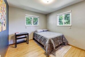 Ein Schlafzimmer mit einem Bett, einem Nachttisch und zwei Fenstern mit Blick auf Bäume.