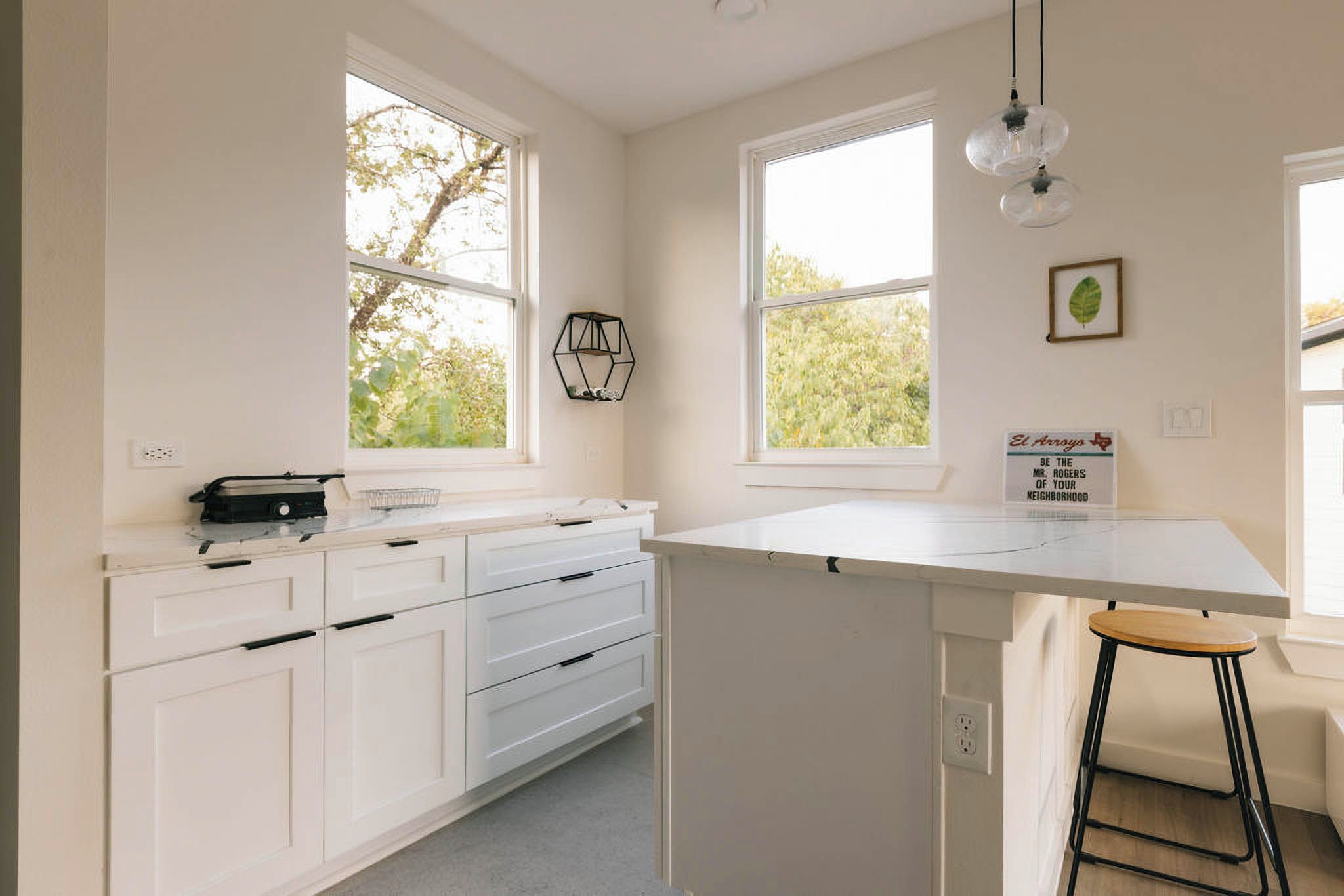 A kitchen with white cabinets, a countertop, and a bar stool. There are two windows with views of greenery outside.