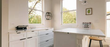 Une cuisine avec des meubles blancs, un plan de travail et un tabouret de bar. Deux fenêtres offrent une vue sur la verdure à l’extérieur.
