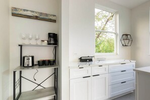 A kitchen with white cabinets, a window with a view of trees, a shelf with kitchenware, and a painting on the wall. - East Austin Tiny Home - 4 Mi to Dtwn! (Austin)
