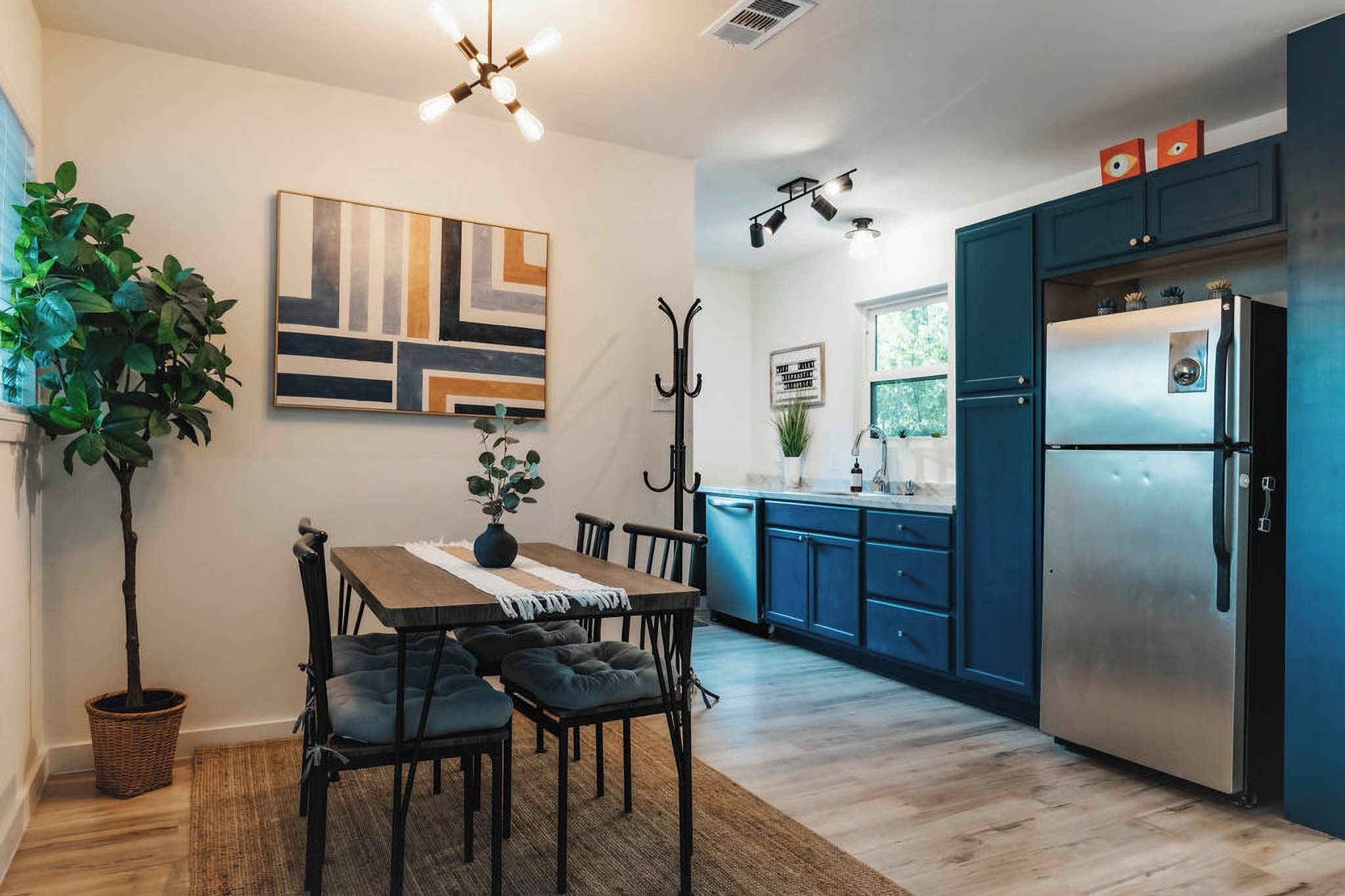 A modern kitchen with a dining area, featuring a stainless steel refrigerator, blue cabinets, a wooden dining table with chairs, and a large potted plant.