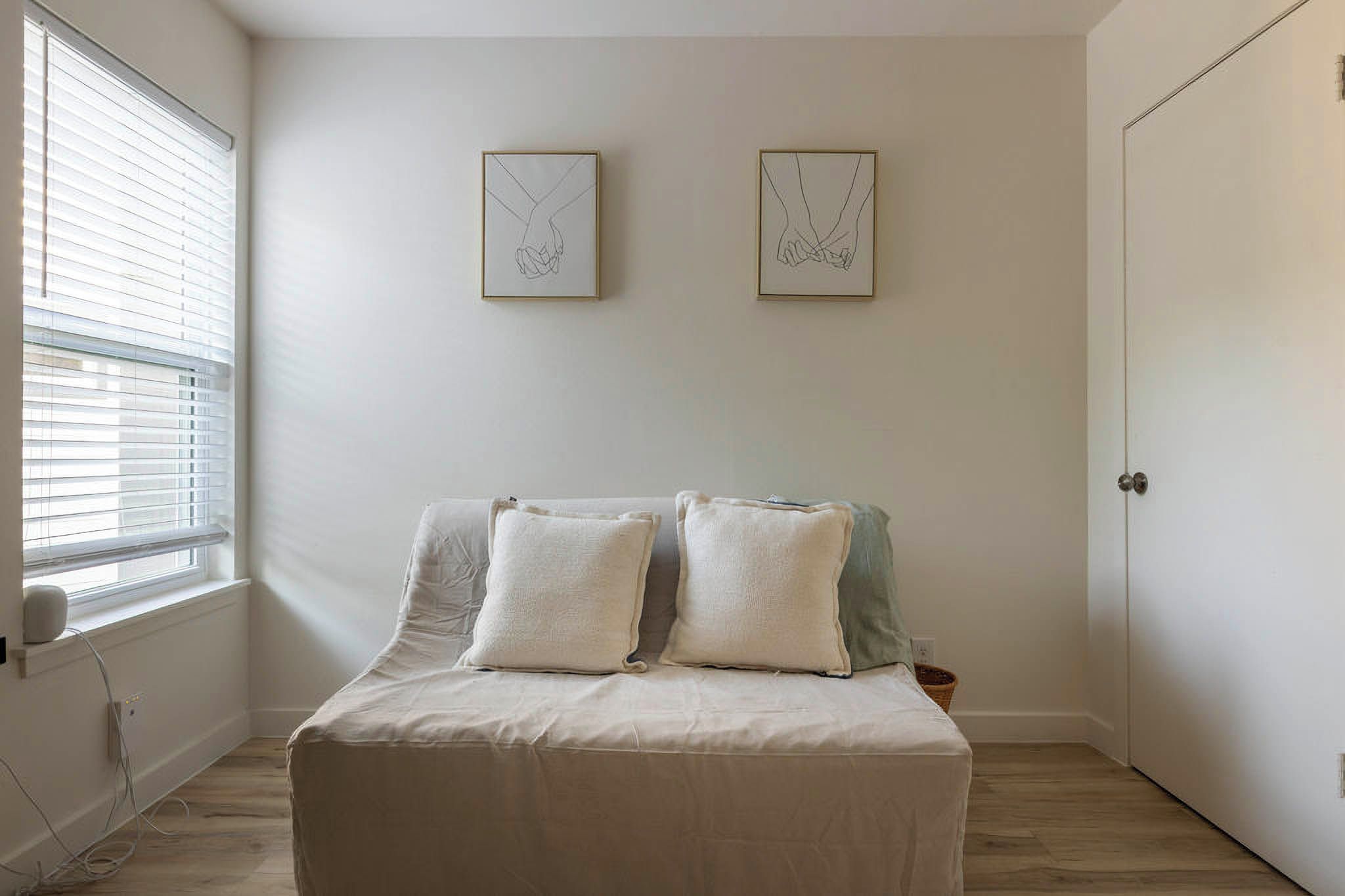 A bedroom with a bed, two pillows, a window with blinds, and two framed artworks on the wall.