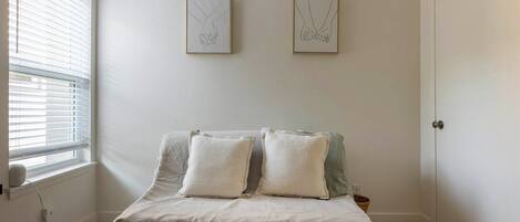 Une chambre à coucher avec un lit, deux oreillers, une fenêtre avec des stores et deux œuvres d’art encadrées accrochées au mur.