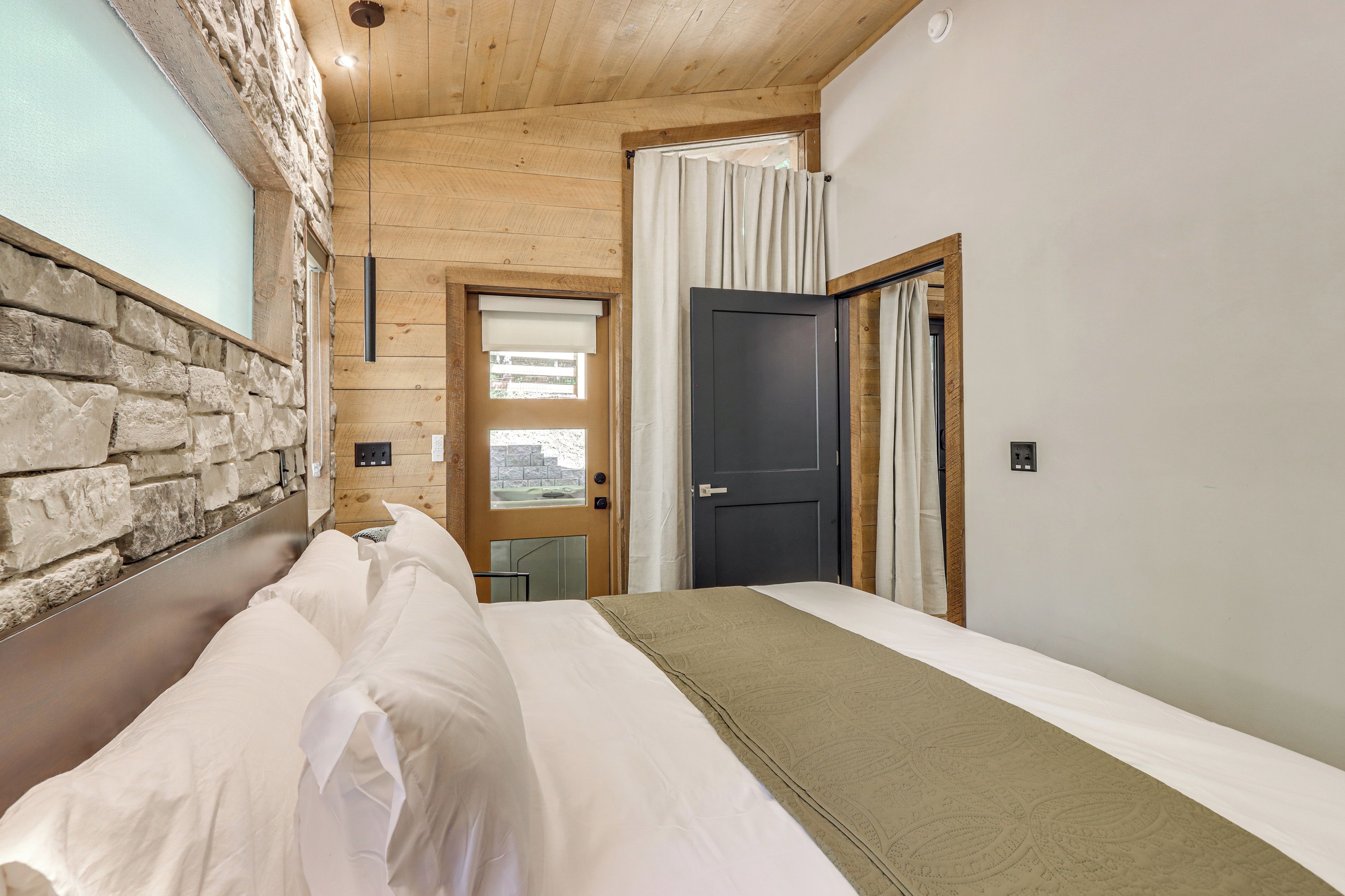 Un dormitorio con pared de piedra, techo de madera, cama con ropa de cama blanca, ventana con cortinas y una puerta.