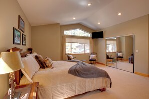 A bedroom with a large bed, a bedside table with a lamp, a chair, a window with blinds, and a mirror on the wall.