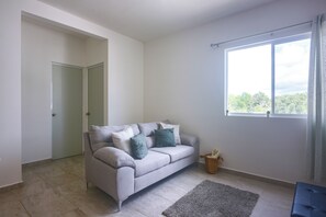 A modern living room with a grey sofa, a small rug, and a window with a view of greenery. - 3 Mi to Beach! Condo w/ Pool in Puerto Morelos (Puerto Morelos)