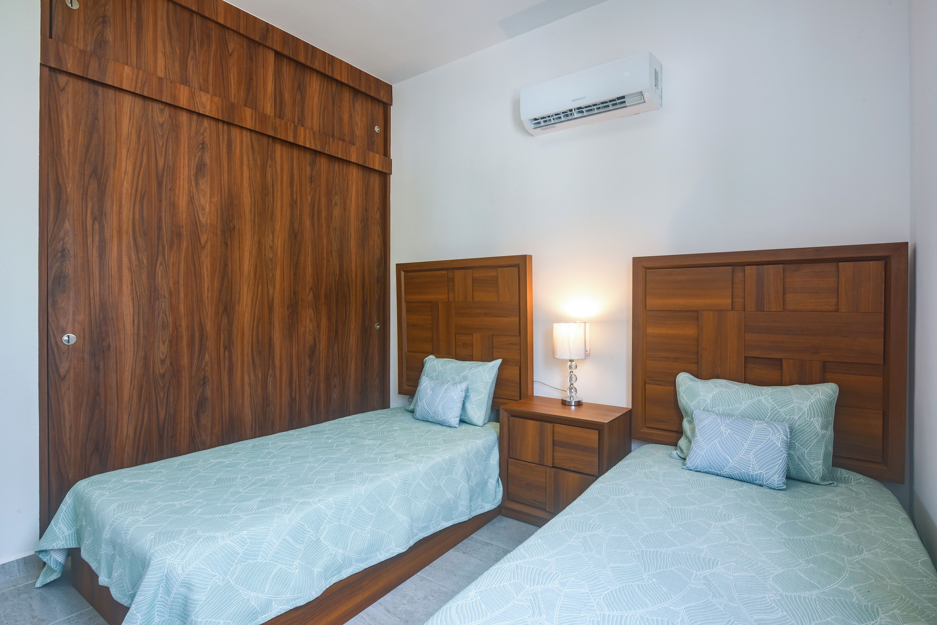 A bedroom with two beds, wooden headboards, a nightstand with a lamp, and an air conditioning unit.