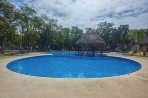Une piscine circulaire, surmontée d’un pavillon au toit de chaume, est entourée de chaises longues et de palmiers.