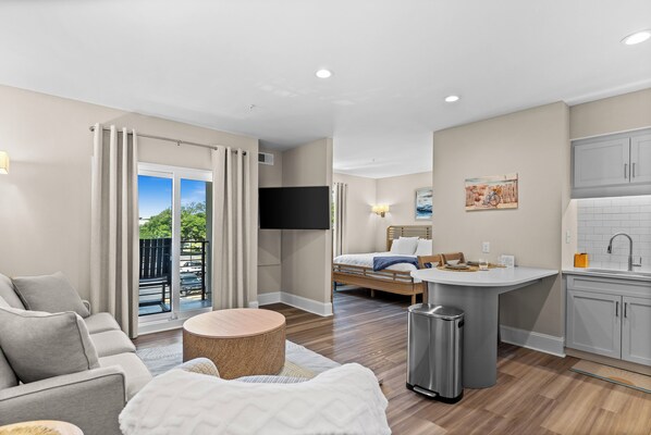 Una habitación de hotel moderna con sofá, una pequeña mesa redonda, cama con cabecera de madera, televisor montado en la pared, zona de cocina con pileta y alacenas, y balcón con vista al estacionamiento.
