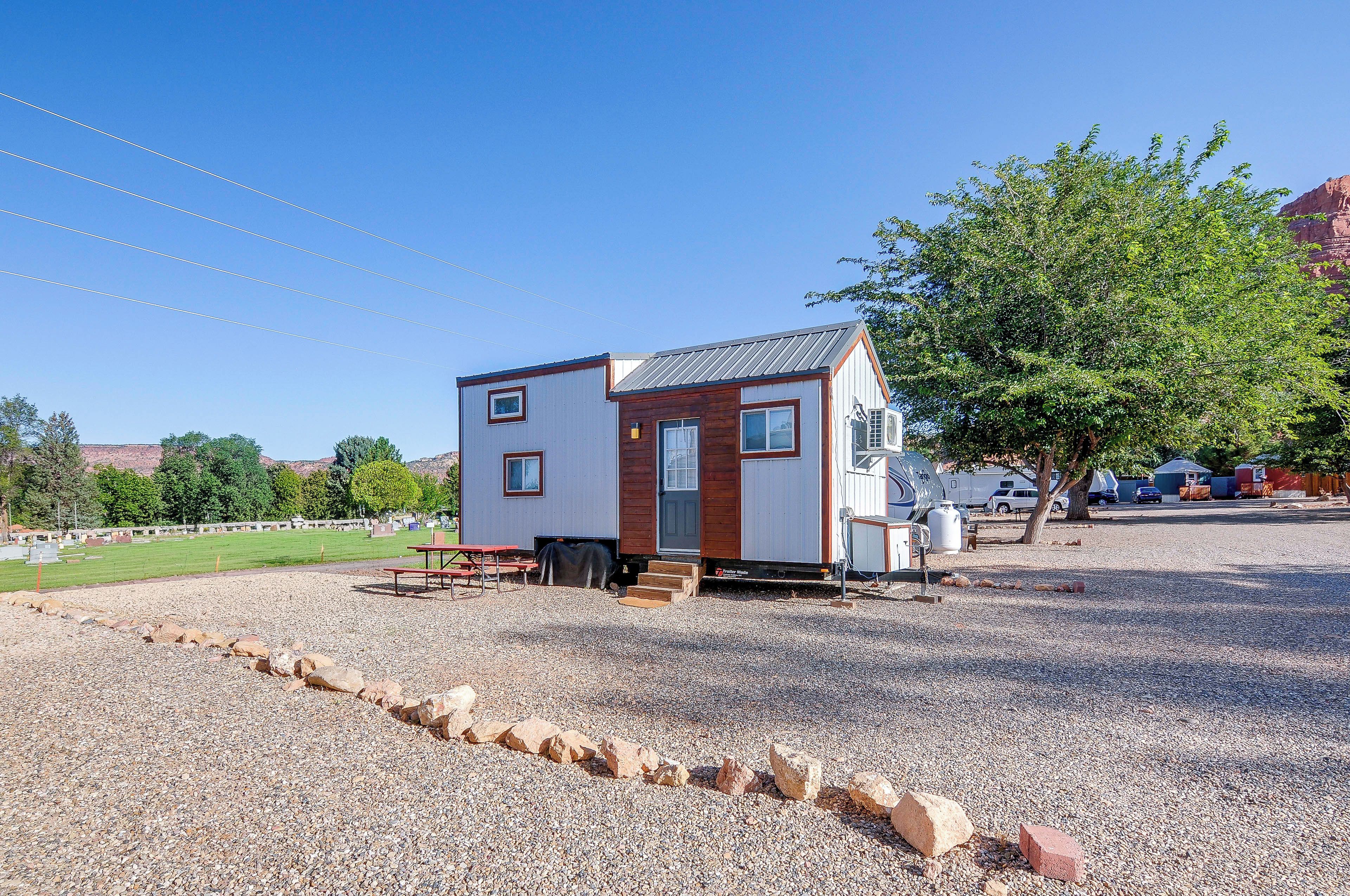 En lille, etetages campingvogn med grusindkørsel, et picnicbord og et træ i baggrunden.