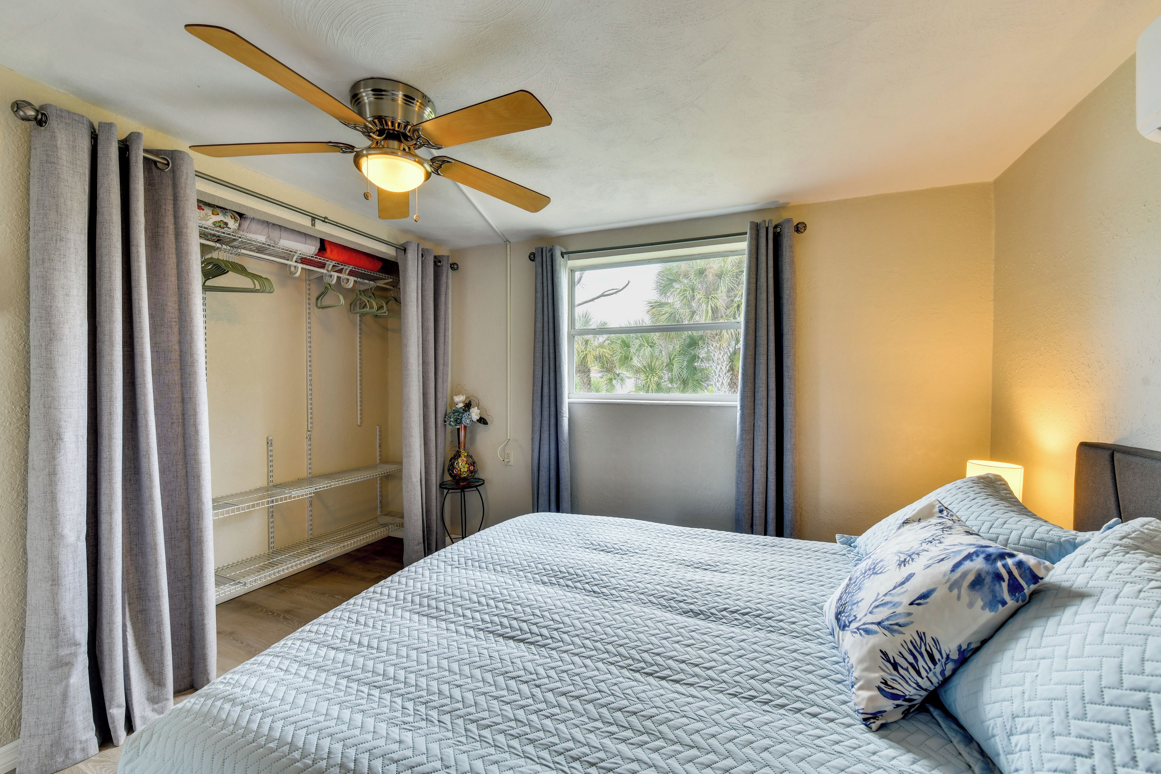 Une chambre à coucher avec un lit, un ventilateur de plafond, une fenêtre et un placard où sont suspendus des vêtements.