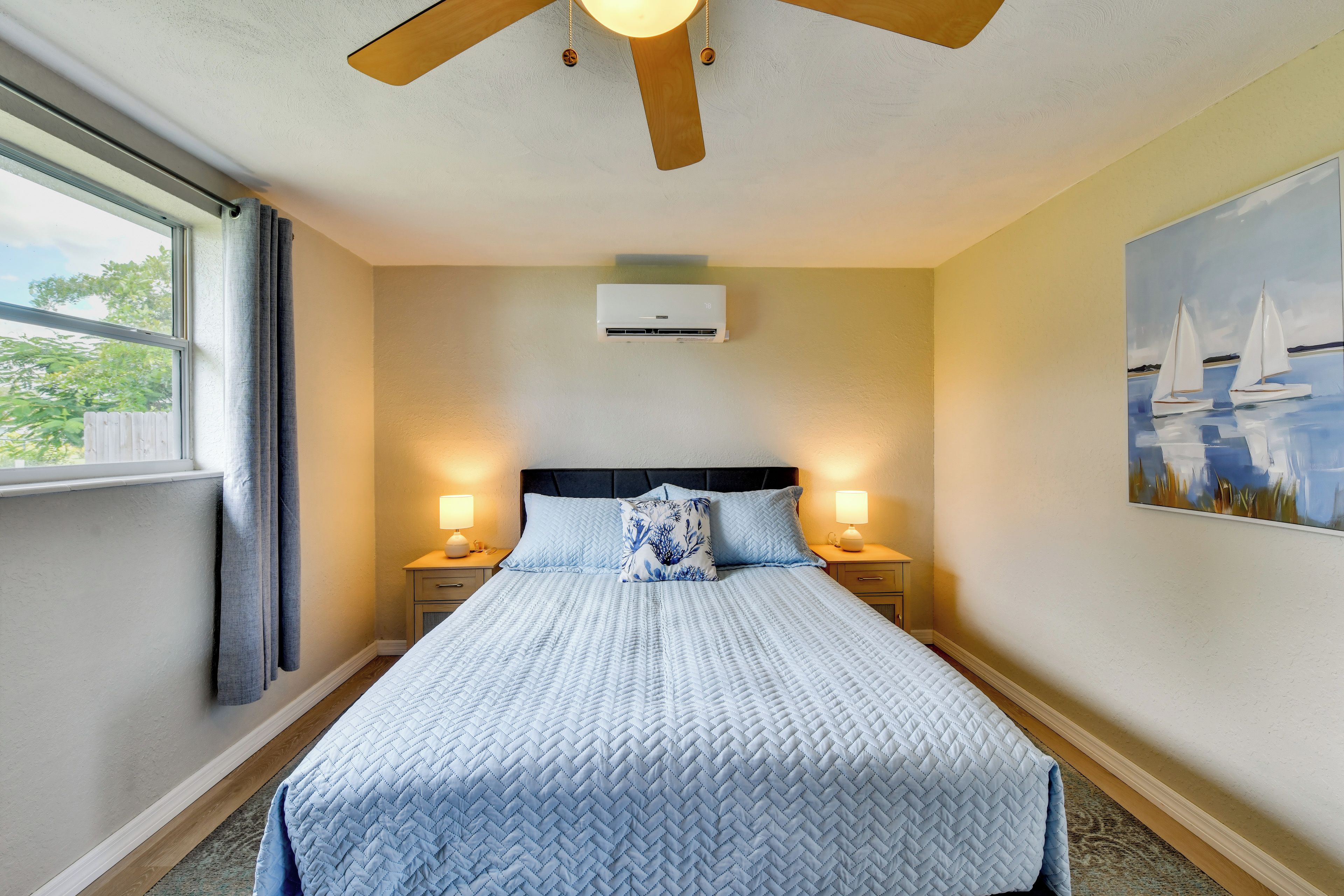 A hotel room with a bed, bedside lamps, an air conditioning unit, a window with curtains, and a painting on the wall.