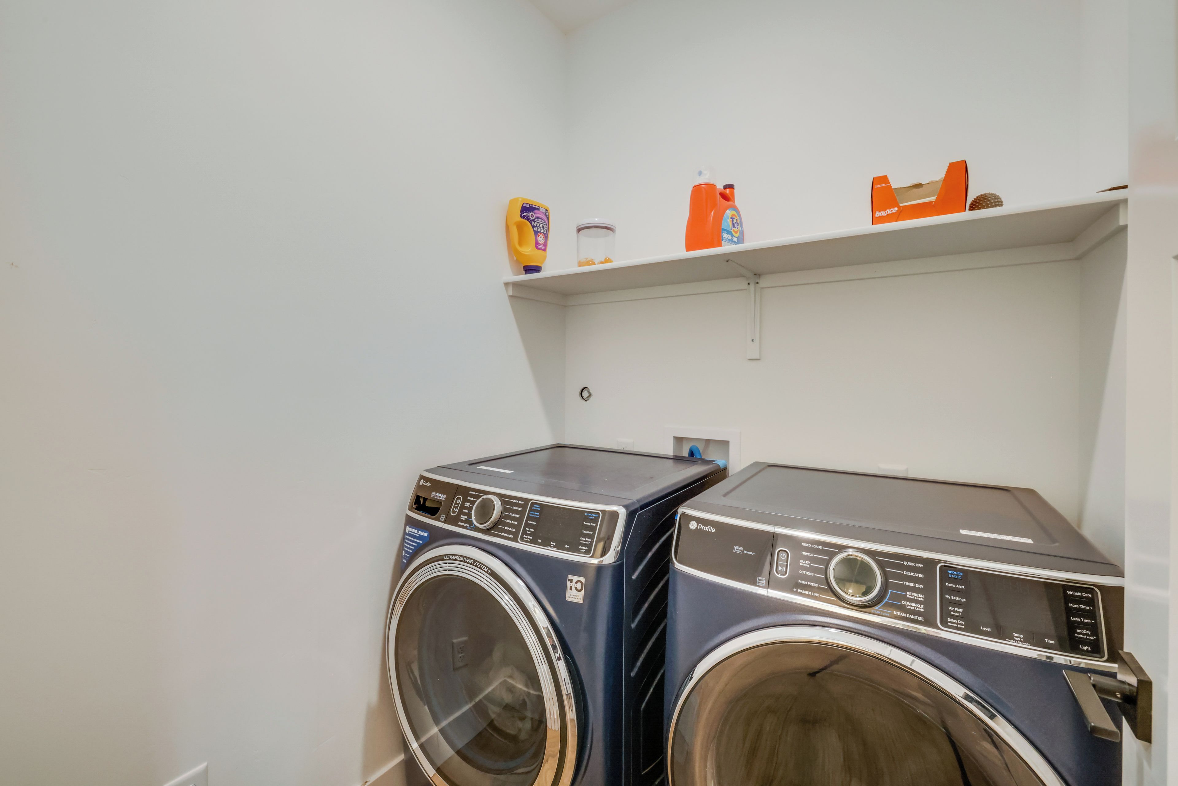 A laundry area with two front-loading washing machines and a shelf above containing cleaning supplies.