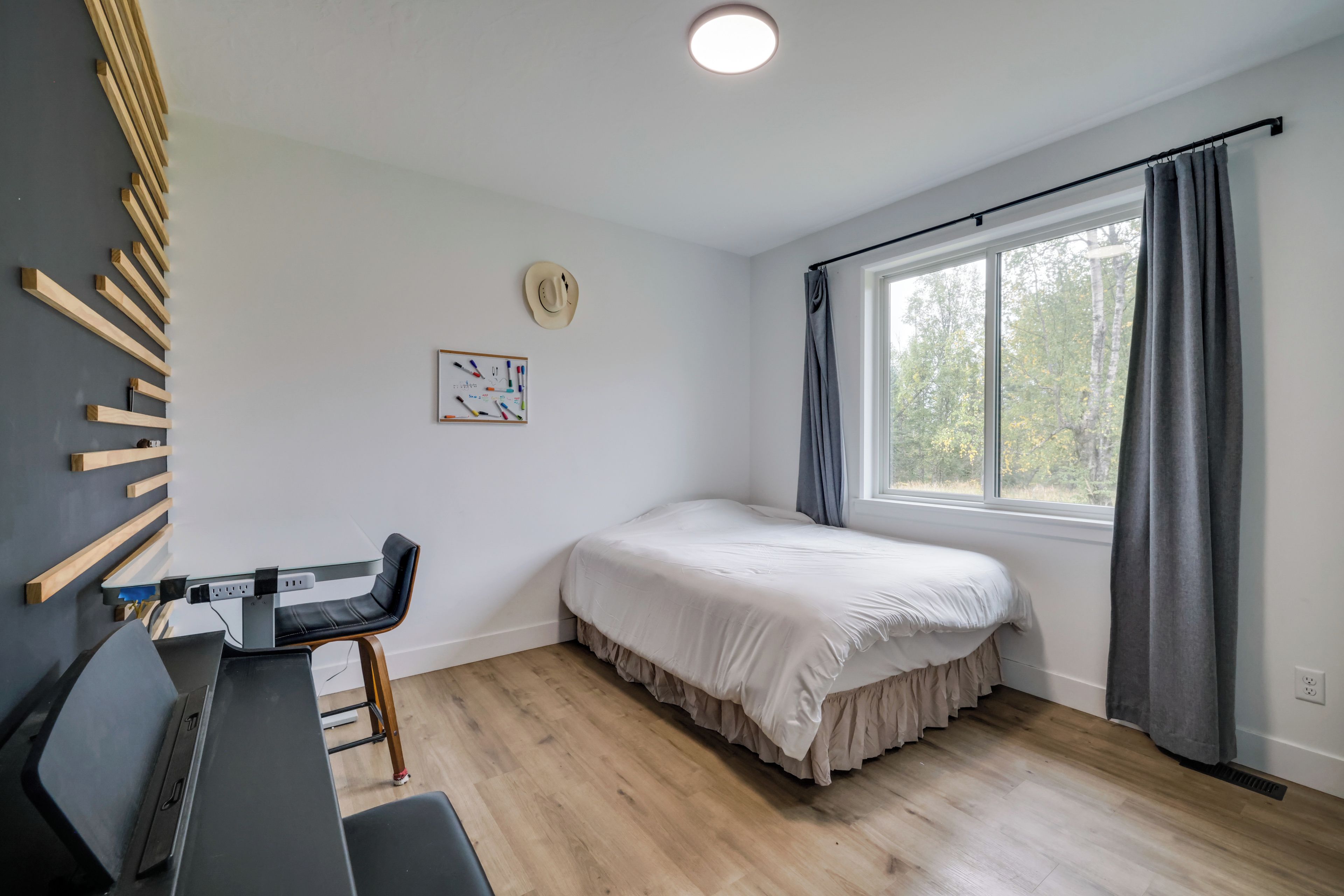 A bedroom with a bed, a desk, a chair, a window with curtains, and a wall-mounted hat.