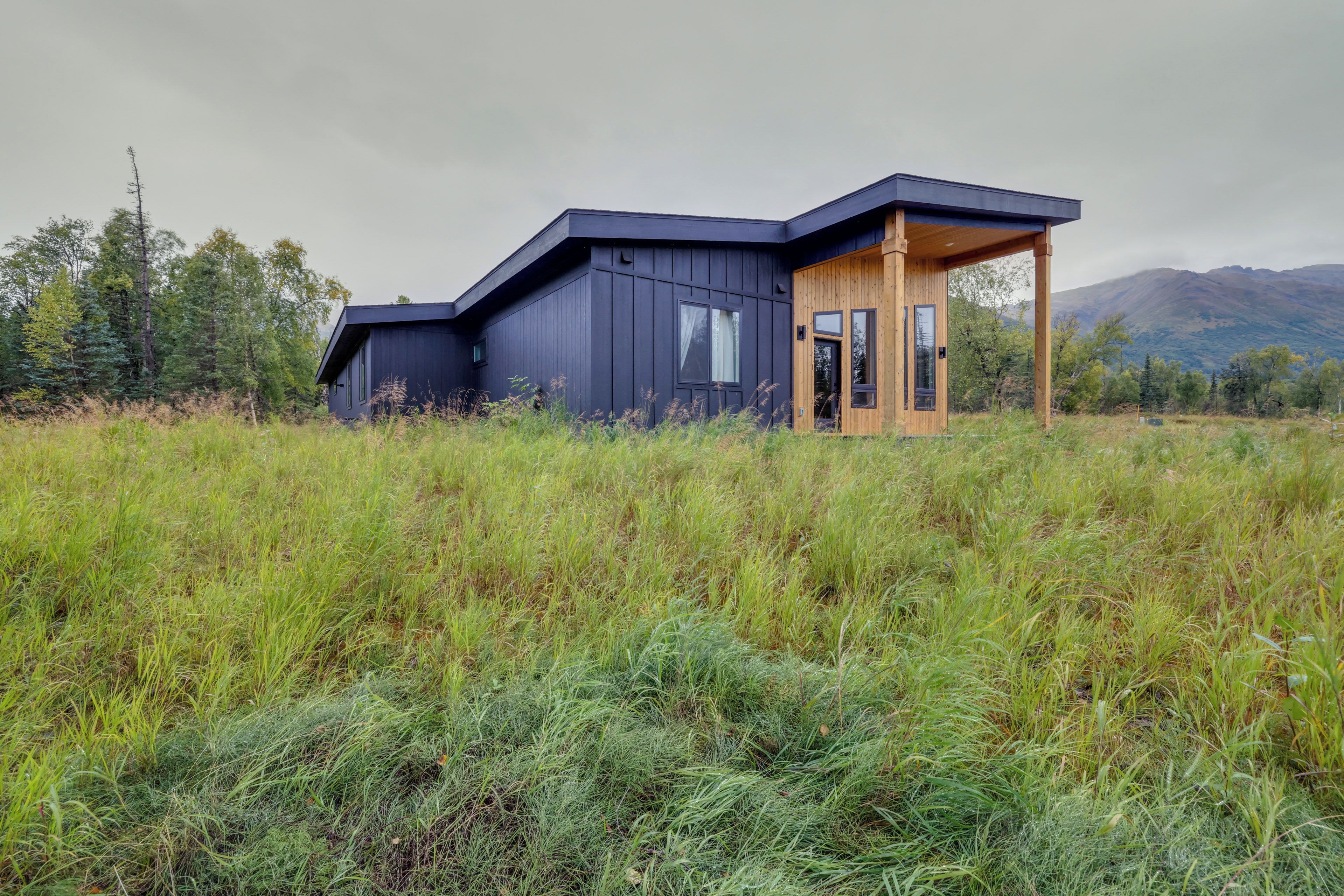 A modern cabin with a dark exterior, a wooden porch, and a view of a mountainous landscape.