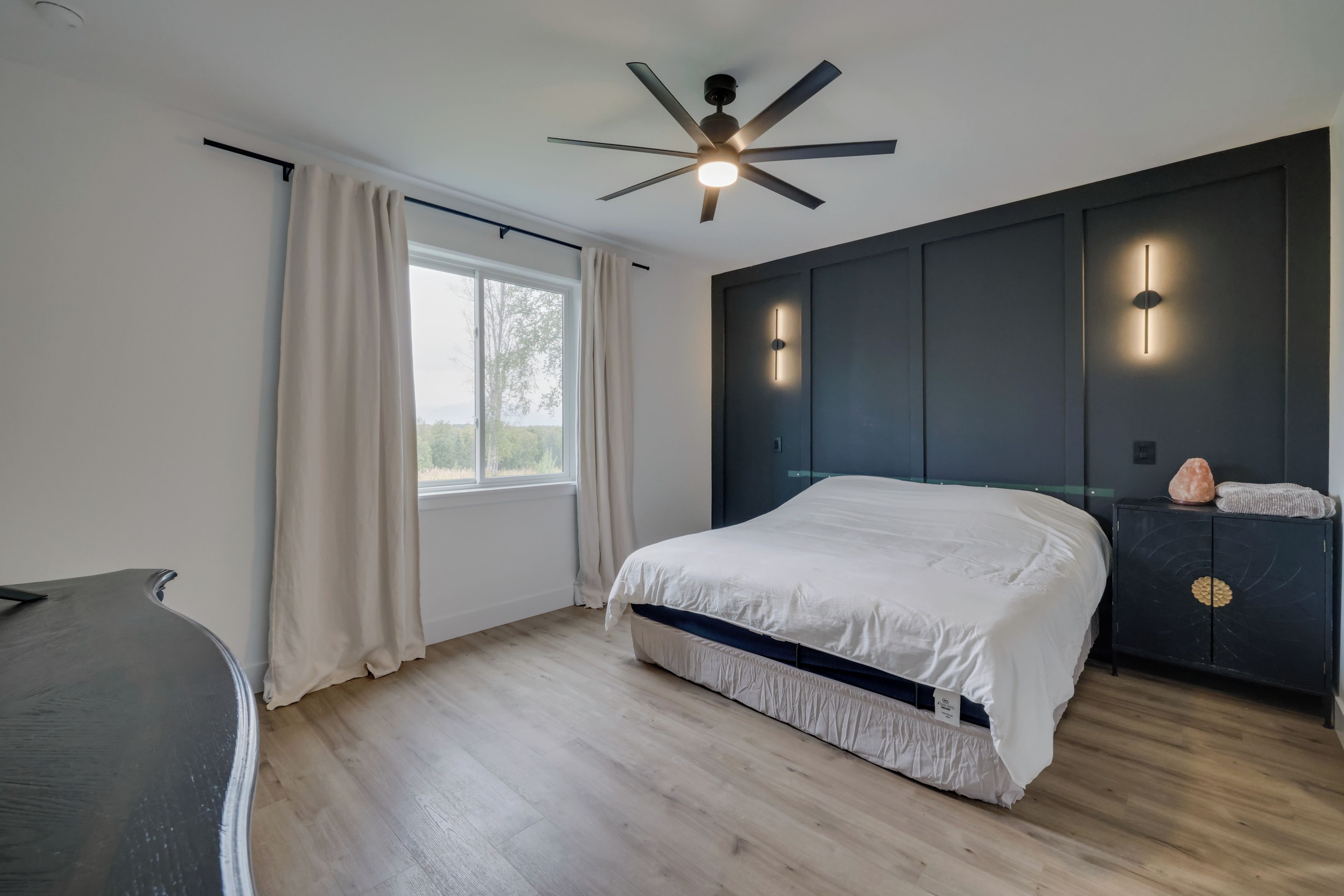 Un dormitorio moderno con cama, ventilador de techo, ventana con cortinas y mesita de noche con lámpara.
