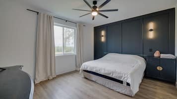 Un dormitorio moderno con cama, ventilador de techo, ventana con cortinas y mesita de noche con lámpara.