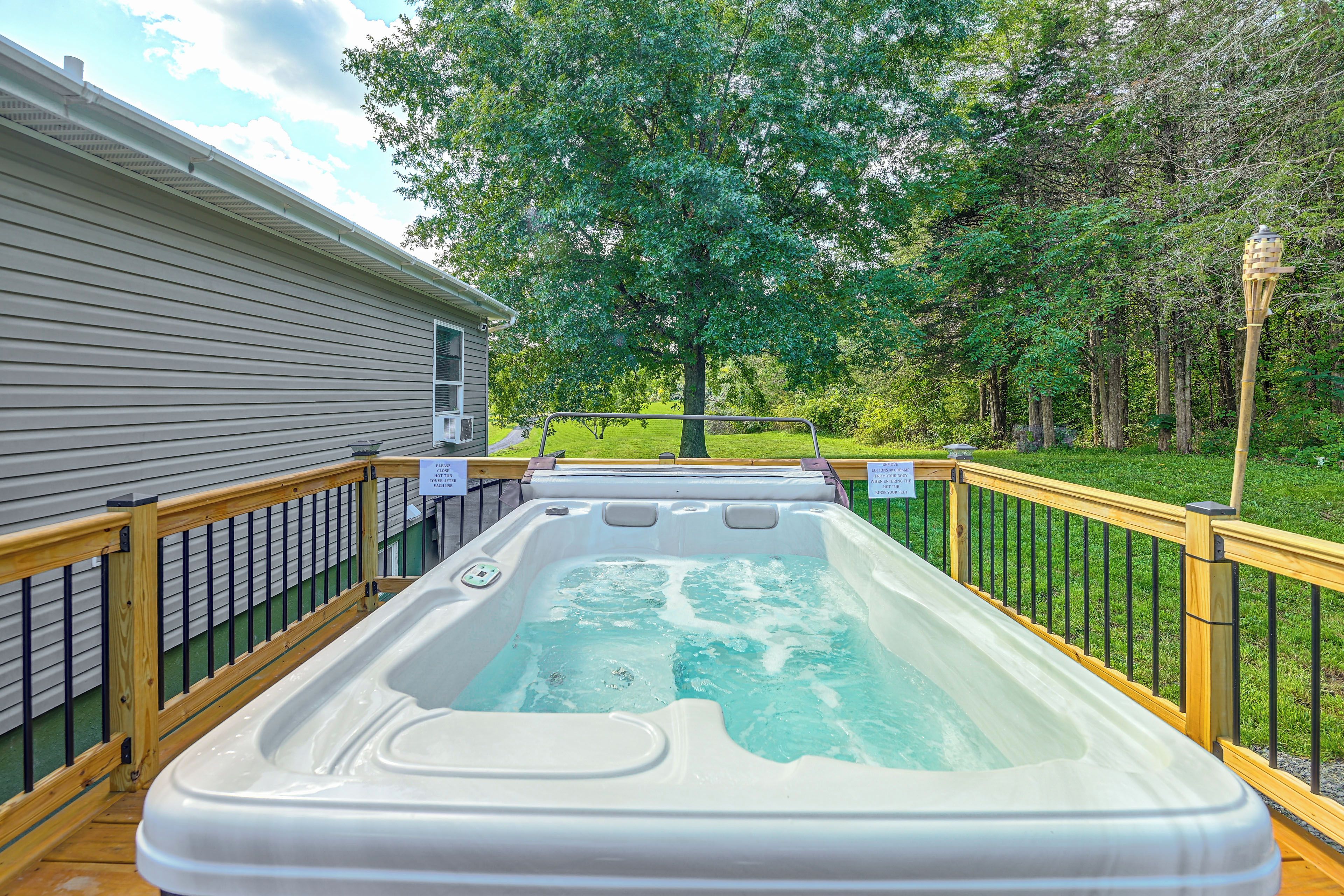 A hot tub on a wooden deck with a metal railing, surrounded by a grassy yard and trees.