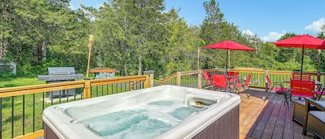 Un jacuzzi en una terraza de madera con un tobogán, rodeado de sillas rojas y sombrillas, y una parrilla en el fondo.