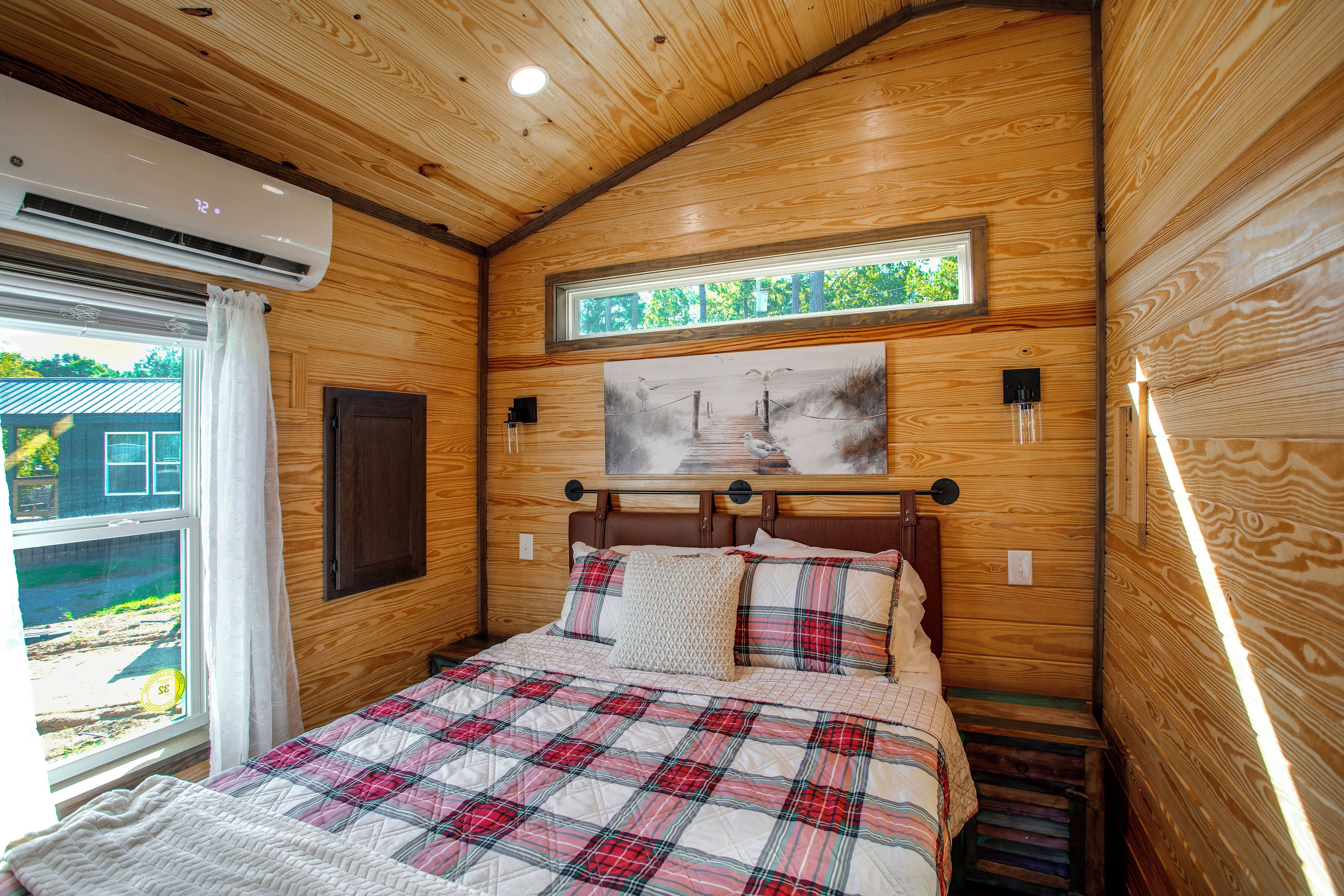 A bedroom with a wooden ceiling, a bed with a plaid quilt, a window with curtains, and a painting above the bed.