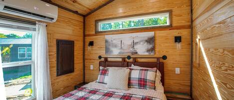 A bedroom with a wooden ceiling, a bed with a plaid quilt, a window with curtains, and a painting above the bed.