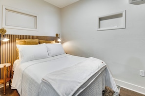 Una cama bien tendida con sábanas blancas, cabecera de madera y mesita de noche con lámpara.