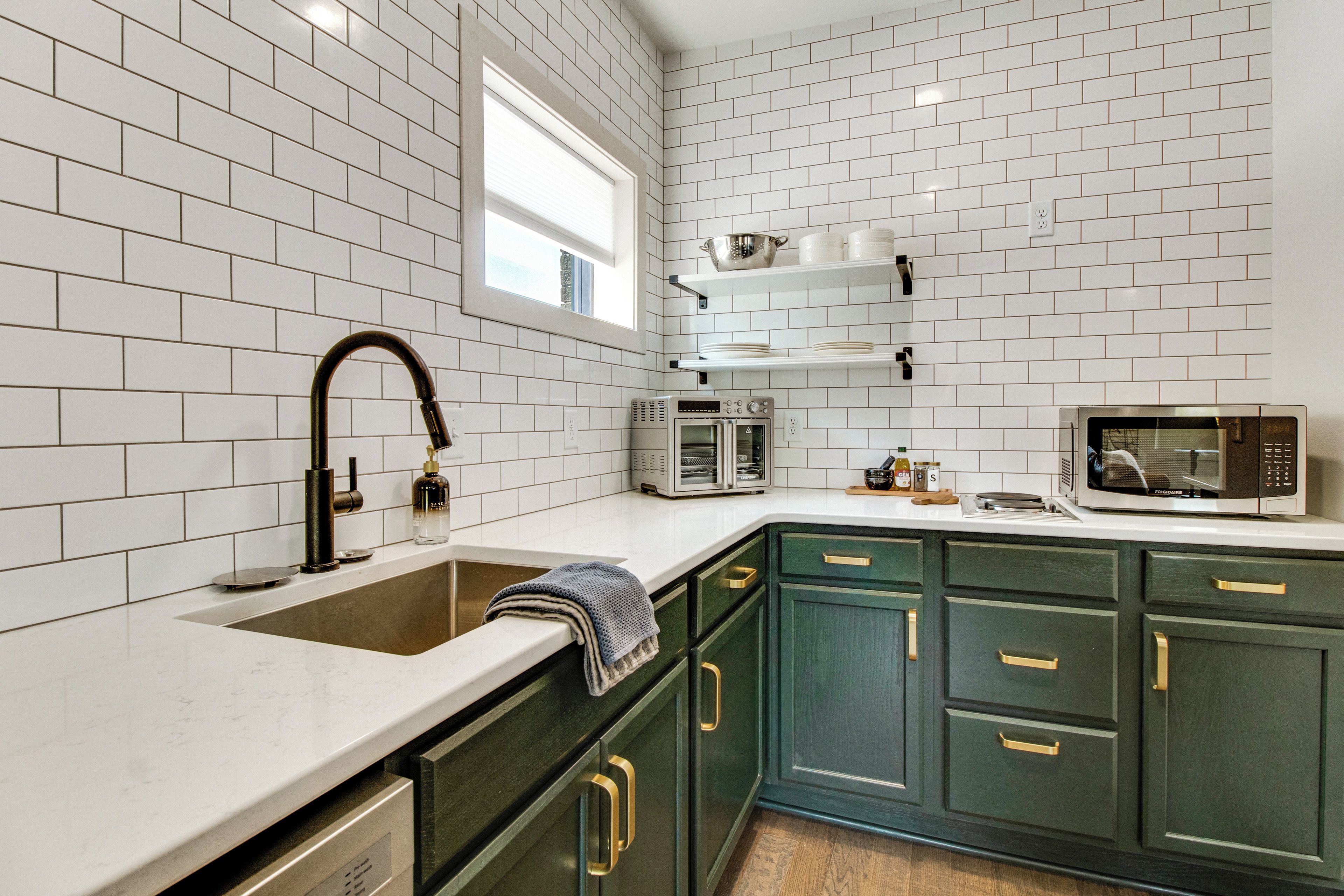 Una cocina con azulejos blancos tipo metro, un lavamanos de acero inoxidable, un microondas y armarios verdes.