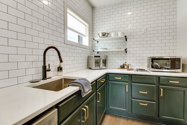 Una cocina con azulejos blancos tipo metro, un lavamanos de acero inoxidable, un microondas y armarios verdes.