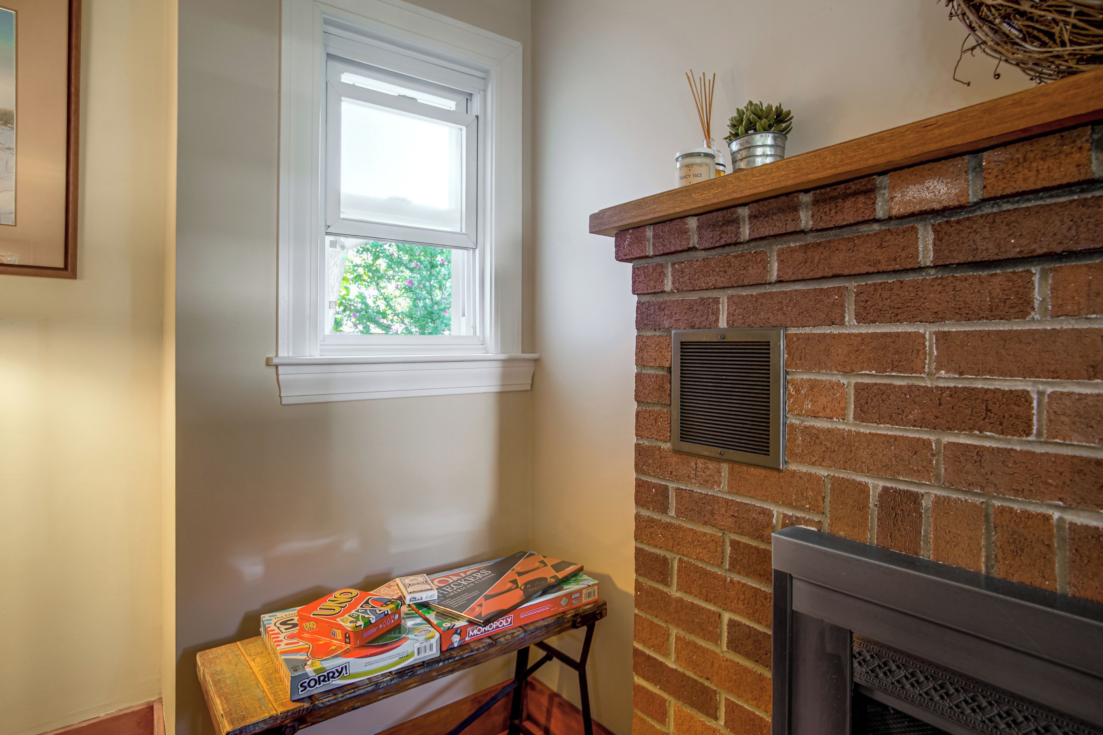 A room with a brick fireplace, a window with a view of greenery, and a table with board games.