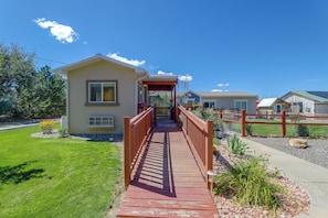 A wooden pathway leading to a single-story house with a covered porch, surrounded by a fenced area and a grassy lawn.