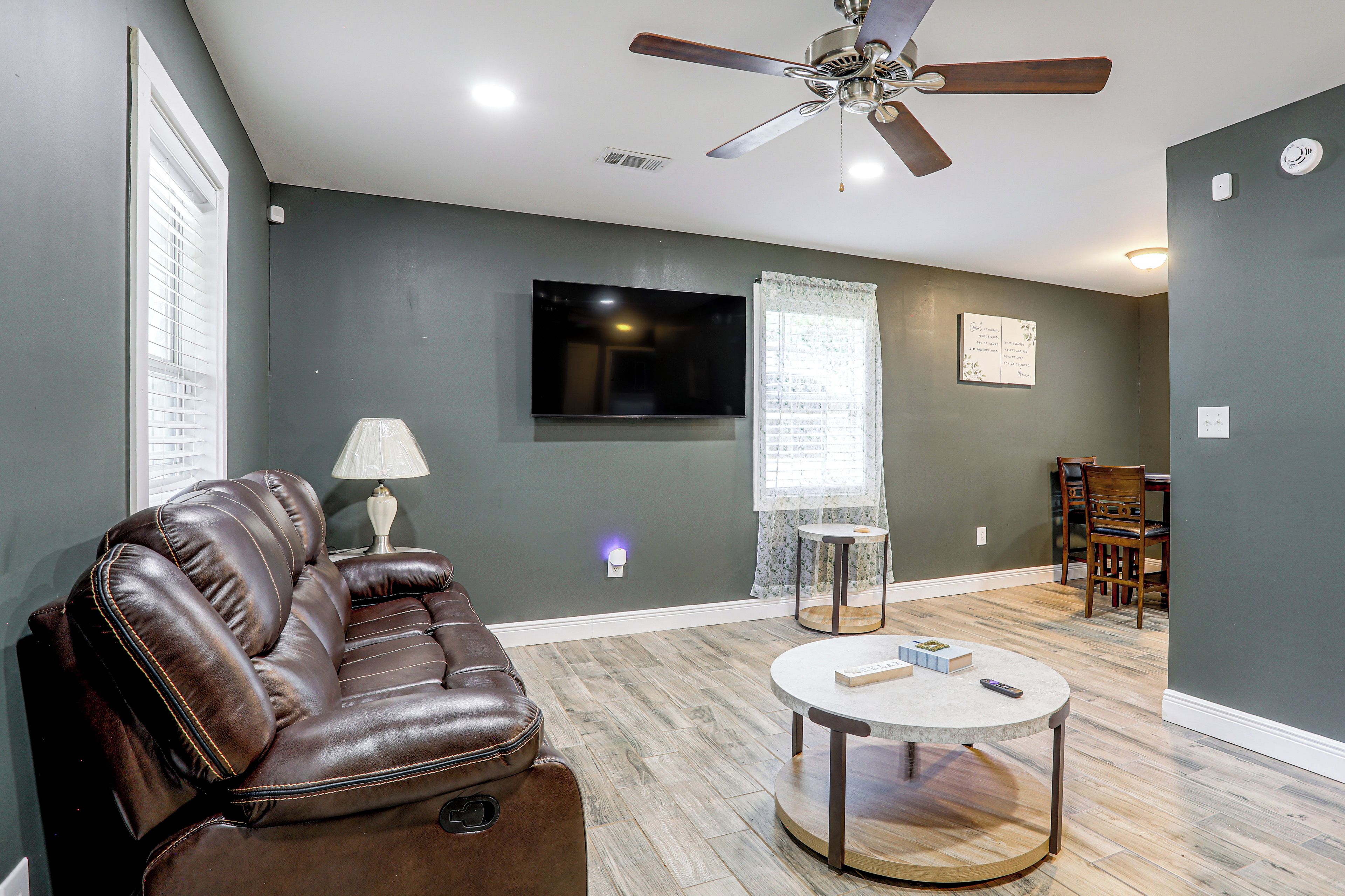 A living room with a leather sofa, a round coffee table, a ceiling fan, a flat-screen TV, and a window with blinds.