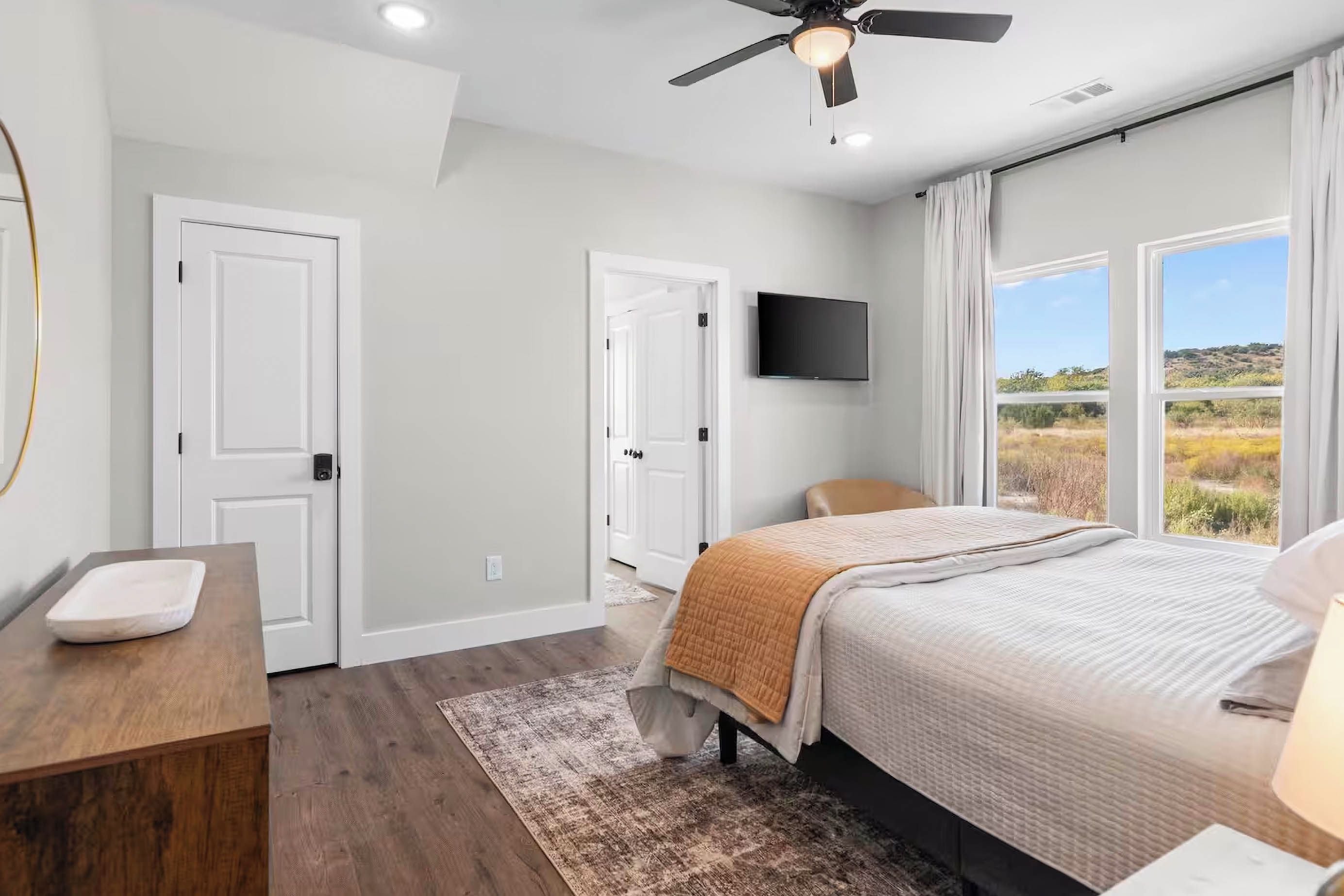 A bedroom with a bed, a desk, a chair, a television, and a view of the outdoors.