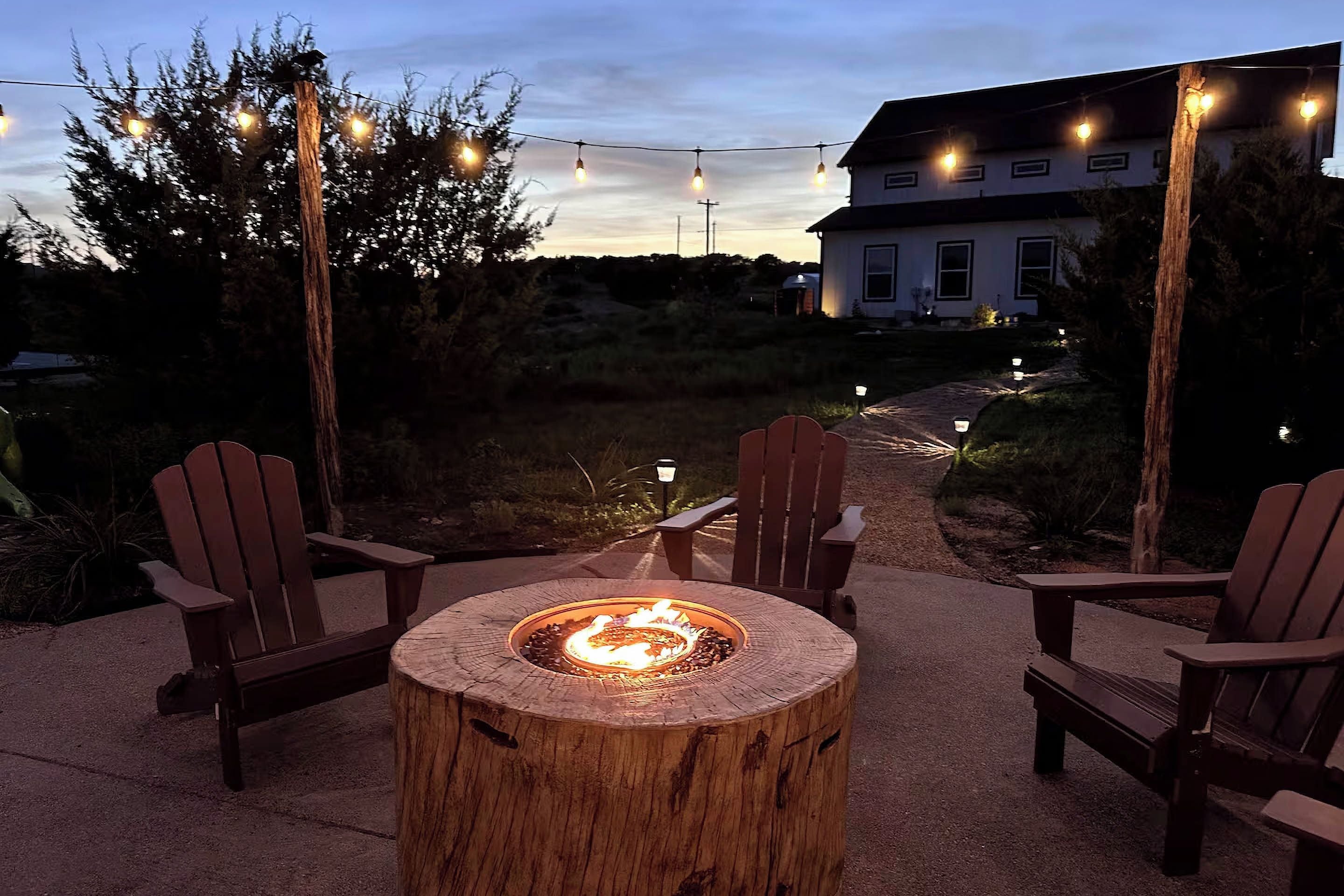 A wooden fire pit with chairs around it, string lights overhead, and a house in the background.
