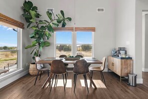 Uma sala de jantar com mesa de madeira, quatro cadeiras, uma grande planta em vaso e vista para o exterior pela janela.