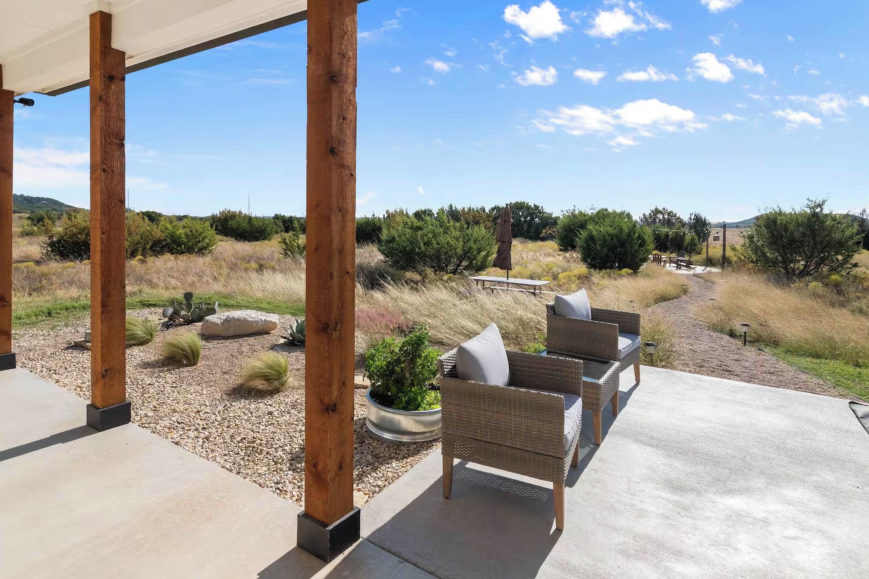 A covered patio with outdoor furniture, a concrete floor, and a view of a natural landscape with shrubs and trees.