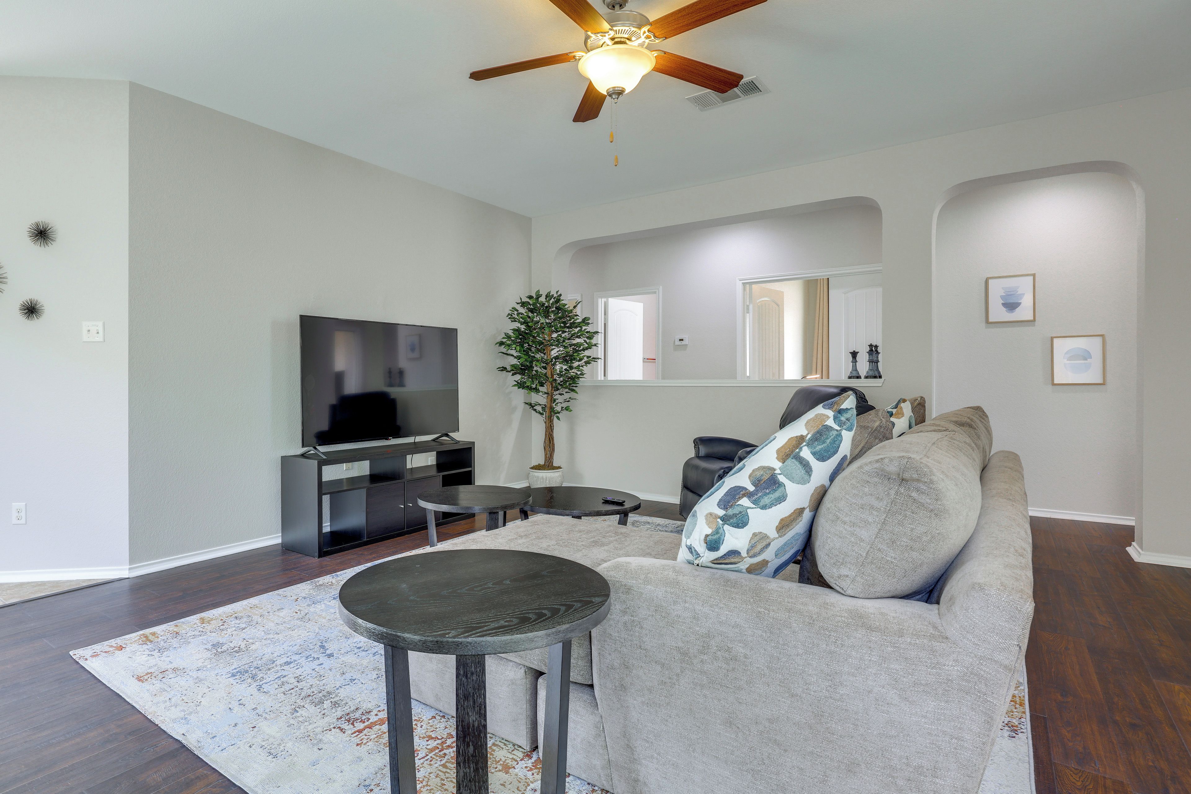 A living room with a flat-screen TV, a ceiling fan, a potted plant, a sofa with patterned cushions, and a coffee table.