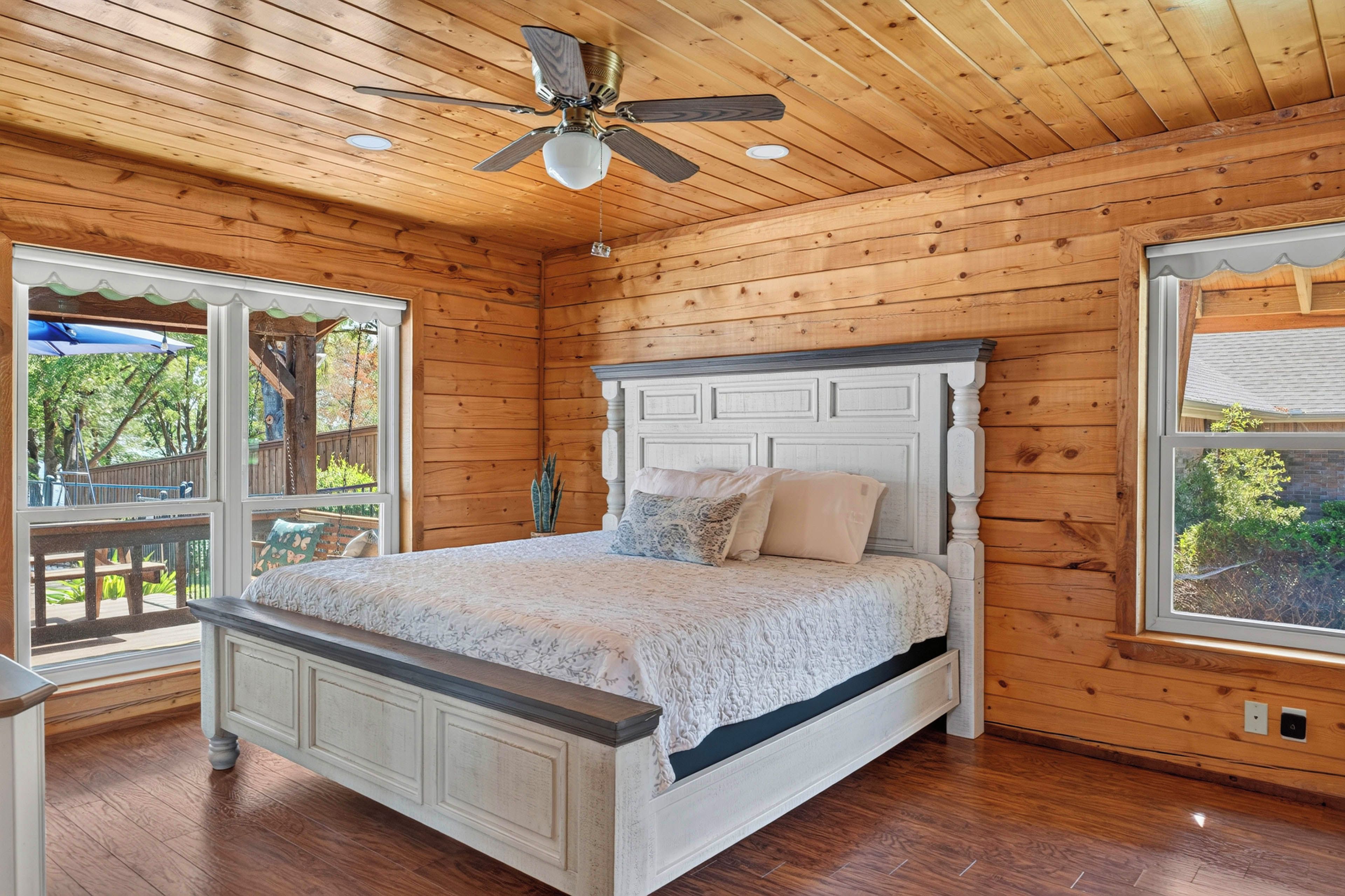 A bedroom with a wooden ceiling, a bed with a white headboard, and large windows.