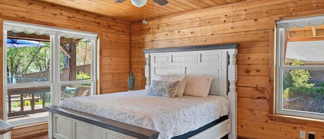 A bedroom with a wooden ceiling, a bed with a white headboard, and large windows.
