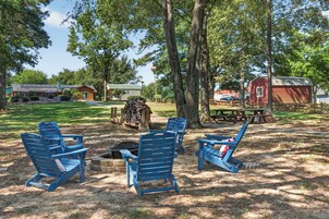 Un camping con sillas Adirondack azules, una fogata, mesas de picnic y un granero rojo al fondo.
