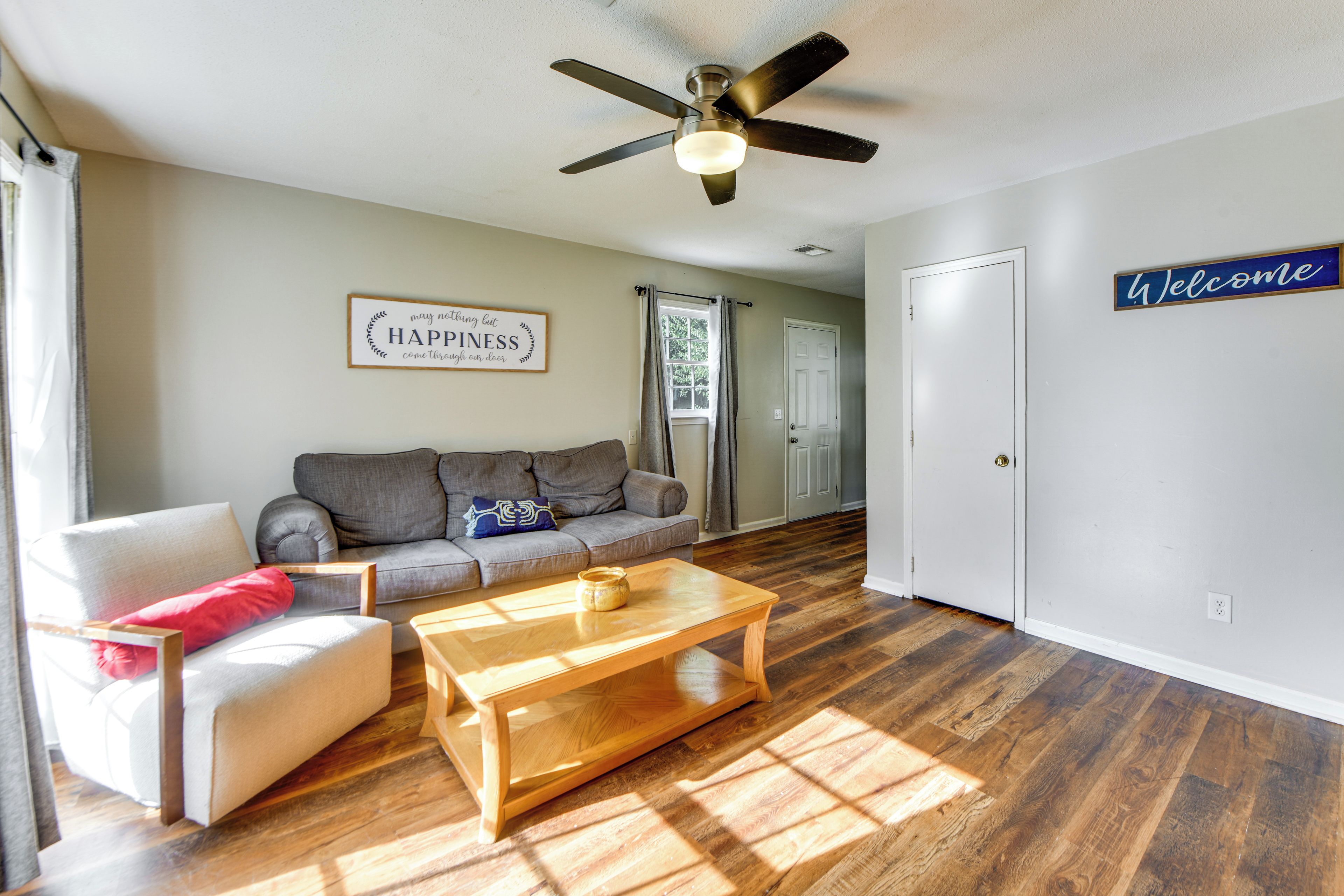 A living room with a sofa, a coffee table, a ceiling fan, and a 'Welcome' sign on the wall.