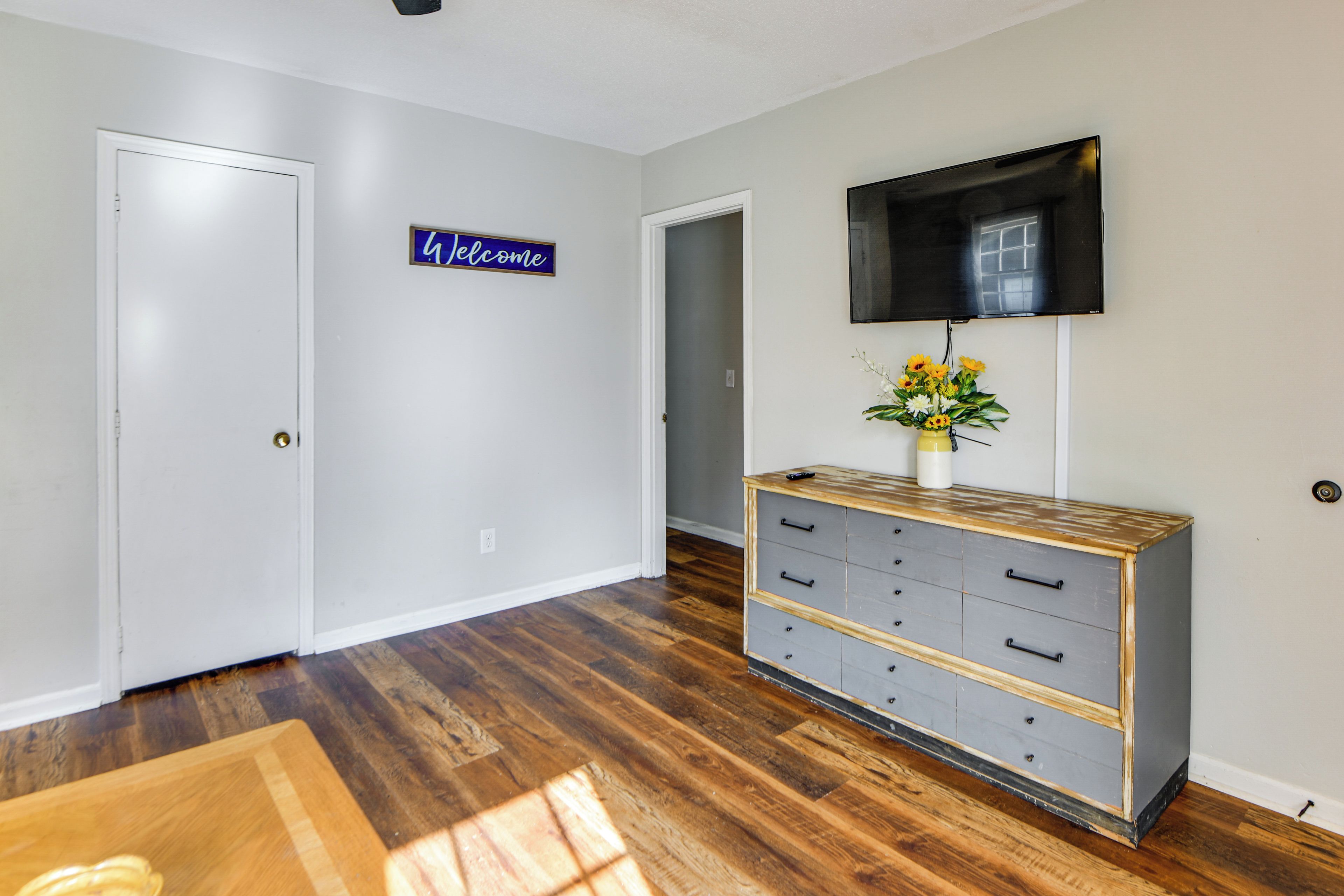 A hallway with a wooden floor, a grey dresser, a TV mounted on the wall, and a 'Welcome' sign.
