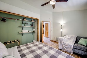 A bedroom with a checkered bedspread, a sofa with a blanket, a wooden shelf with decorative items, and a ceiling fan.