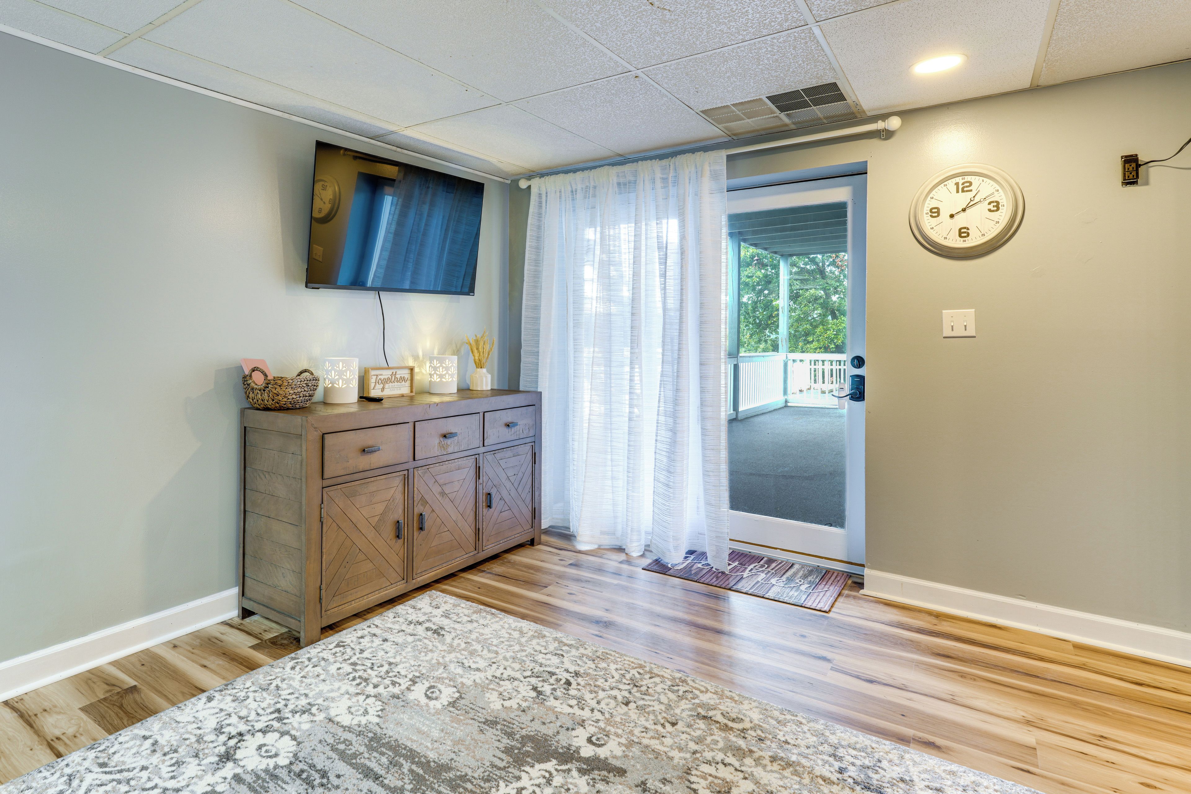 A room with a wooden cabinet, a television mounted on the wall, a patterned rug, and a door with a glass panel.