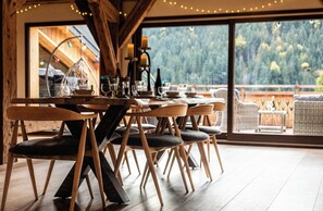 Un coin repas avec des chaises en bois et une table dressée pour quatre personnes, de grandes fenêtres donnant sur la verdure et des guirlandes lumineuses suspendues au plafond.