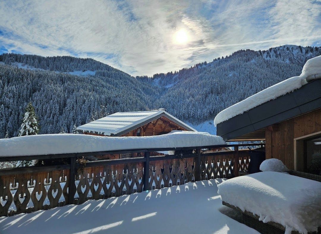 Eine Holzhütte mit schneebedecktem Dach, umgeben von einem Holzzaun, vor einer Kulisse aus schneebedeckten Bergen und einem klaren Himmel.