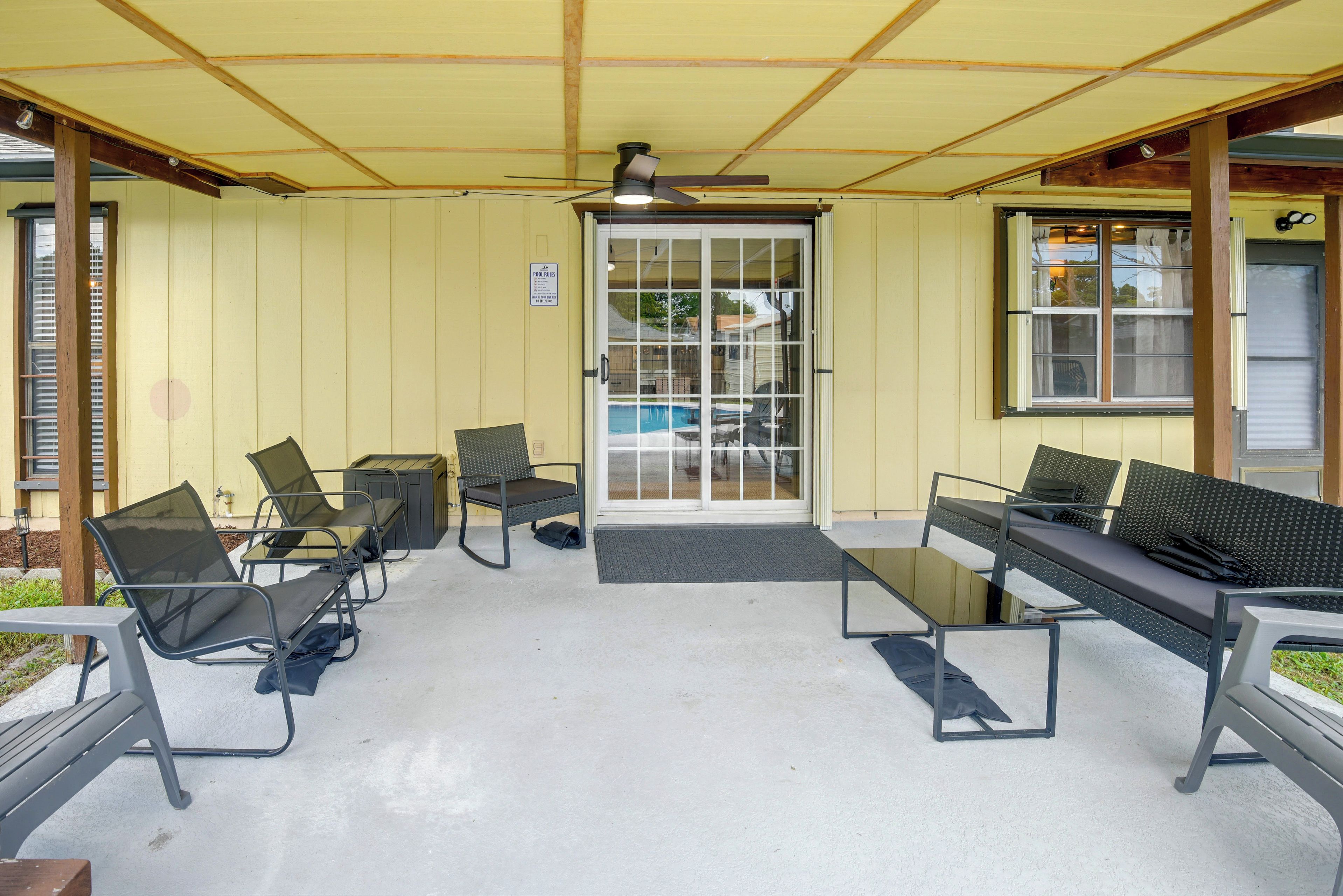 Uma área de estar coberta ao ar livre com cadeiras e uma mesa, piscina visível através de uma porta de vidro e um ventilador de teto.